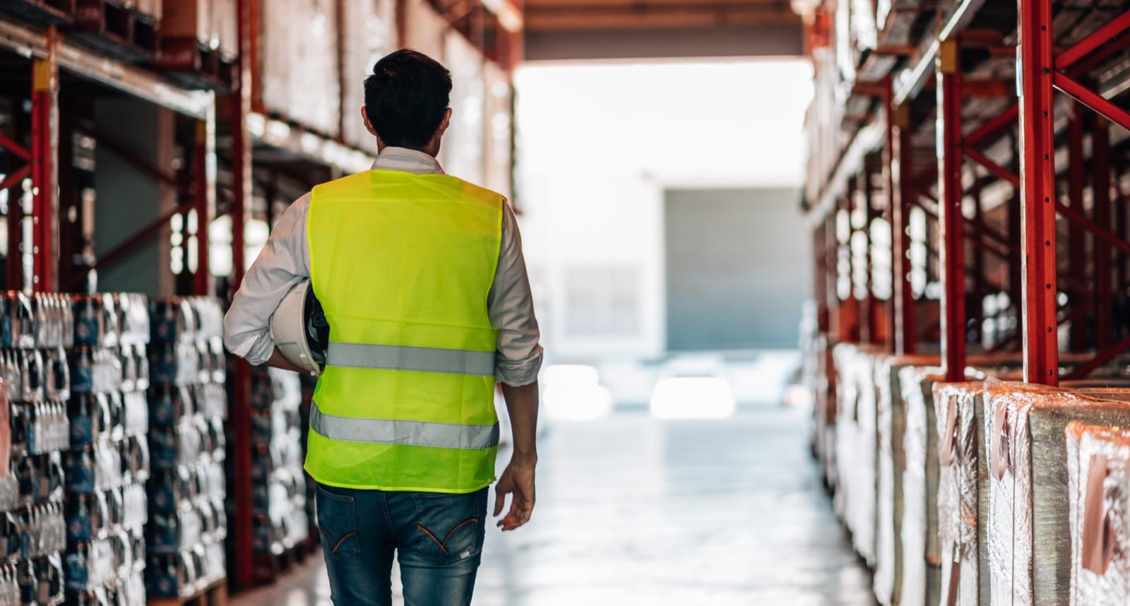 Warehouse worker seen from behind, walking down a storage aisle while holding a hard hat.